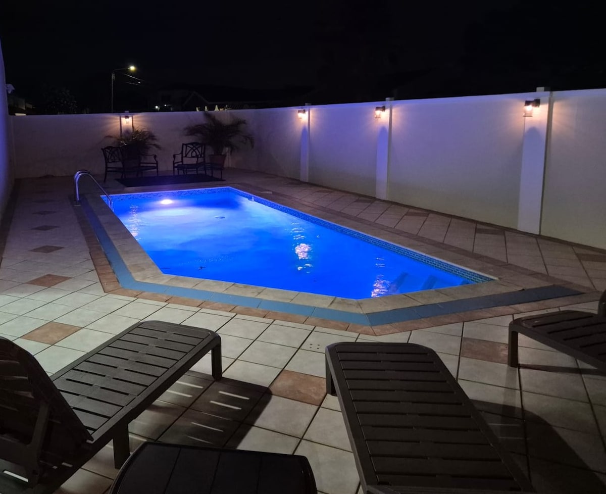 A softly lit pool area is showcased, with a rectangular pool illuminated in shades of blue. The surrounding patio features several lounge chairs, and the walls are adorned with wall-mounted lights, creating a calm nighttime ambiance.