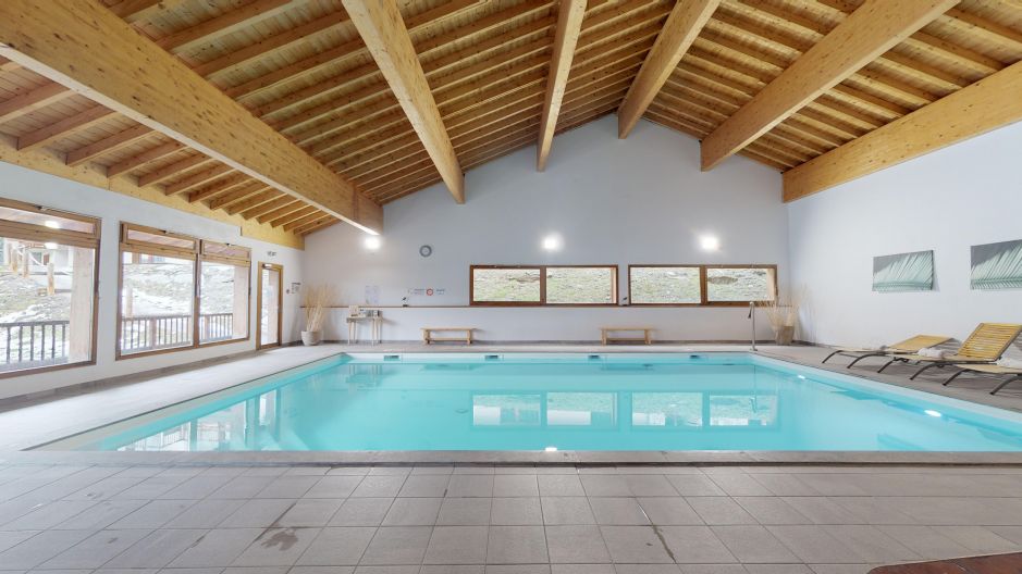 An indoor swimming pool is featured under a high wooden ceiling with exposed beams. Large windows offer natural light and views of the surroundings. Loungers are positioned along the poolside, providing a space for relaxation. The area is clean and inviting.