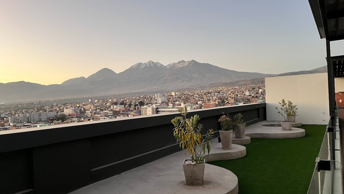 A terrace is showcased featuring a grassy area and several potted plants. In the background, the silhouette of mountains is visible against a soft evening sky, while the cityscape stretches below, highlighting urban life.