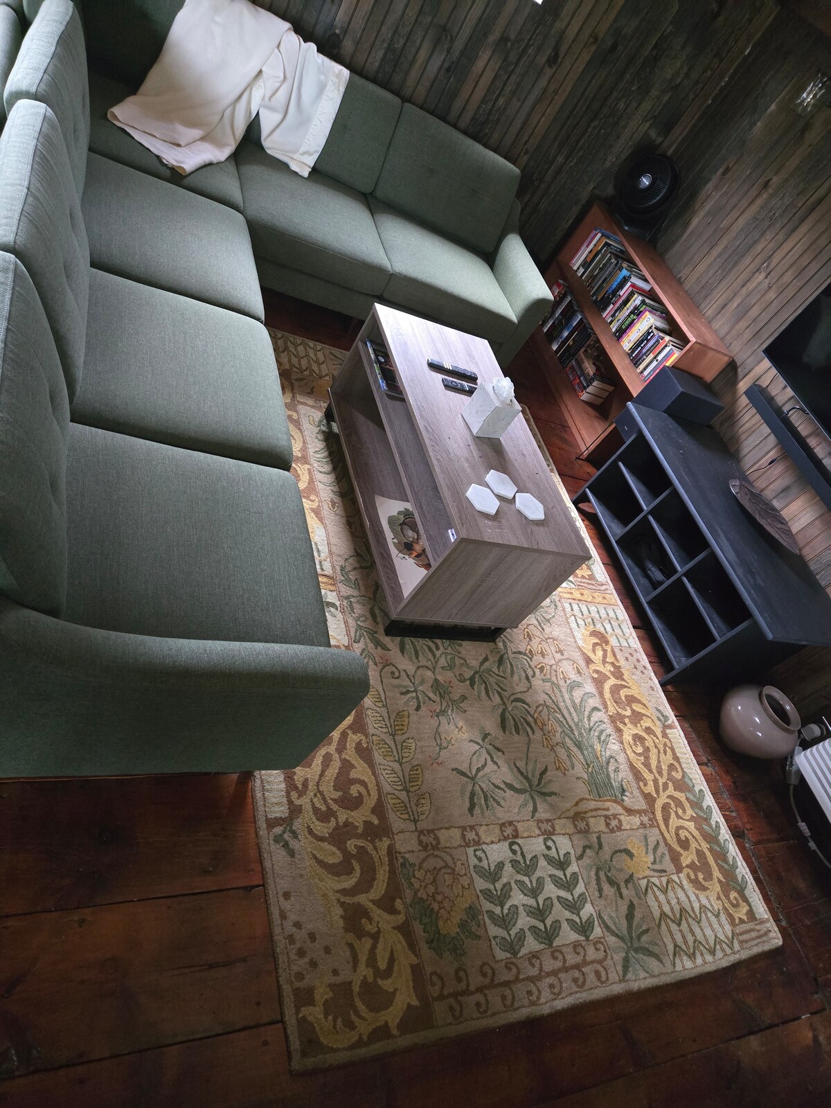 A cozy living area is presented with a green sectional sofa arranged around a wooden coffee table. A patterned area rug adds warmth to the space, while a bookshelf is visible in the background, filled with various books. Soft lighting is provided by a nearby lamp.