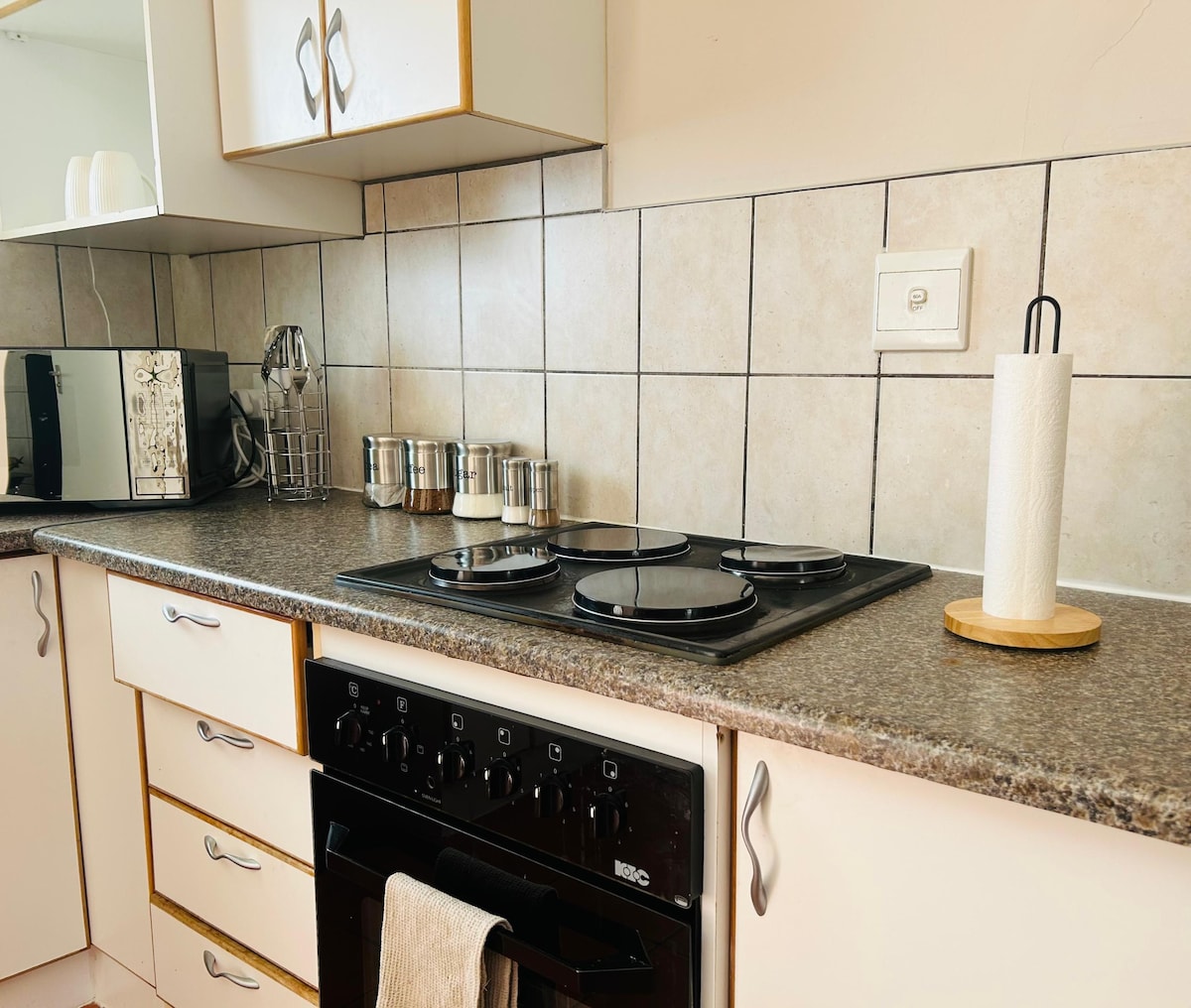 A well-equipped kitchen is presented, featuring a stove with four burners, an oven, and a microwave. The countertops are neatly organized with stainless steel containers and a paper towel holder, while cabinets provide ample storage space. The tile backsplash adds a clean and modern touch.