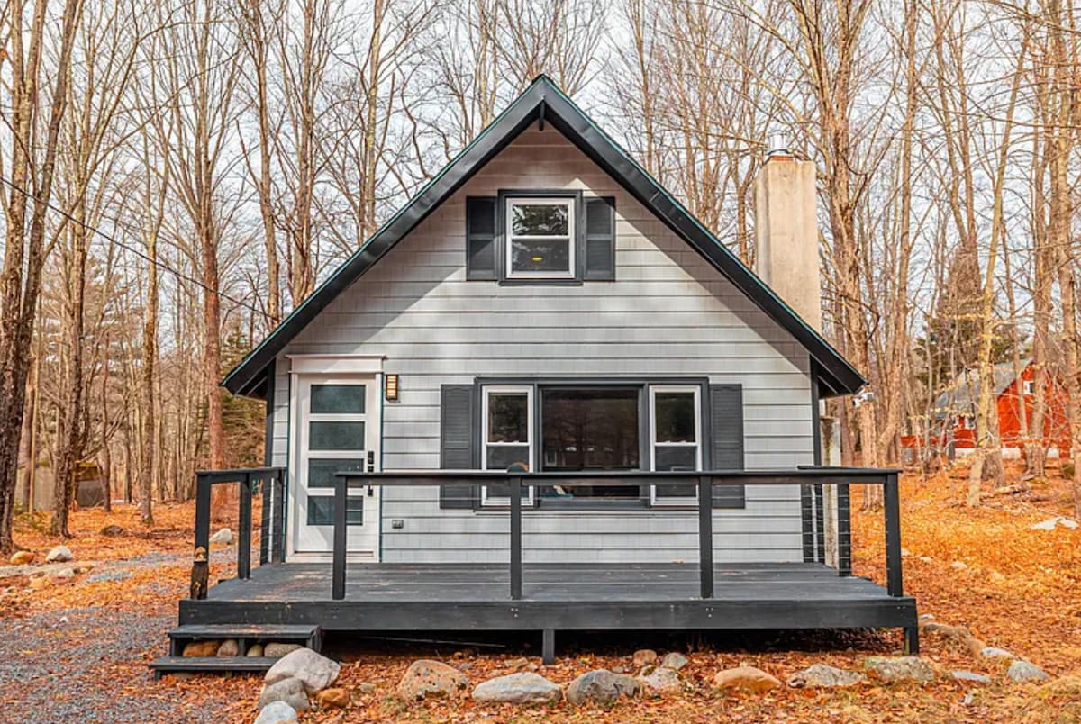 A charming a-frame house is set within a wooded area, surrounded by autumn foliage. A welcoming front deck with a black railing is visible, leading to a door framed by large windows. The natural landscape enhances the home's inviting presence.