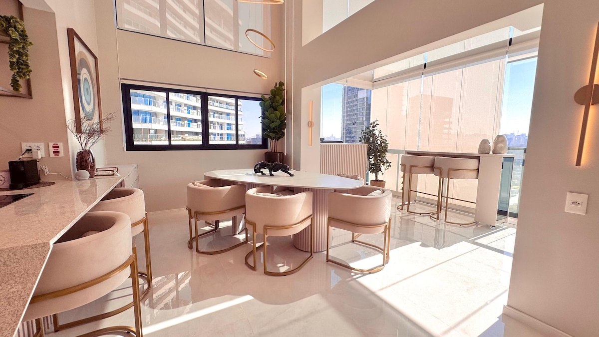 A modern dining area is presented with a circular table, surrounded by soft, upholstered chairs. Large windows allow ample natural light to fill the space, showcasing a serene view. A small plant adds a touch of greenery, complementing the light-colored walls and sleek furnishings.