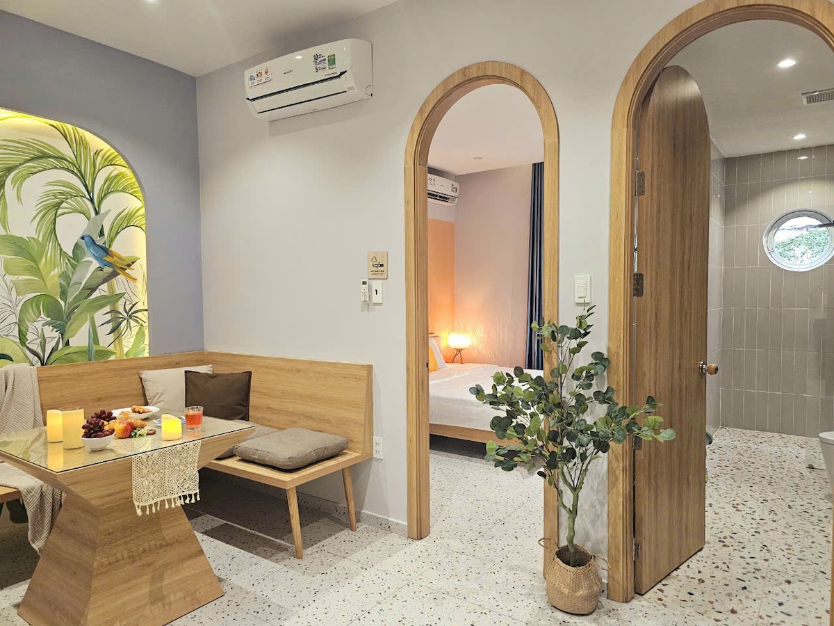 An open space is featured, showcasing a wooden dining table surrounded by benches. A plant adds a touch of greenery. Arched doorways lead to a serene sleeping area with soft lighting, while a circular window allows natural light into the bathroom area.