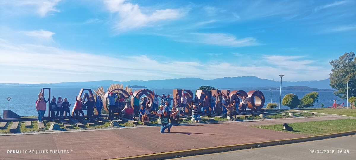 A large sign reading 'Lago Ranco' is prominently displayed, with people posing around it. The blue lake and distant mountains form a scenic backdrop. Sunlight brightens the scene, highlighting the grassy area and walkway in the foreground.