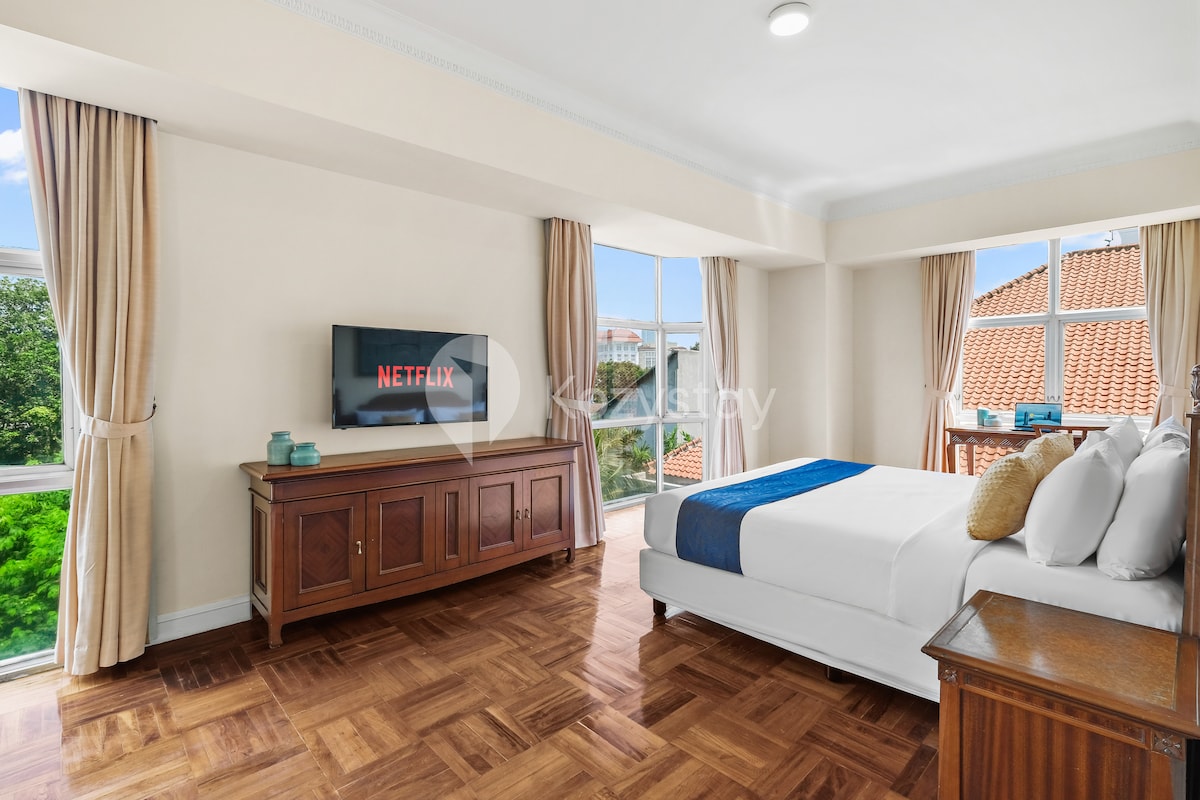 A spacious bedroom features two large windows allowing natural light to flood the area. A comfortable bed with crisp linens is complemented by an attractive wooden nightstand and dresser. A 43'' Smart TV is mounted on the wall, displaying the Netflix logo.