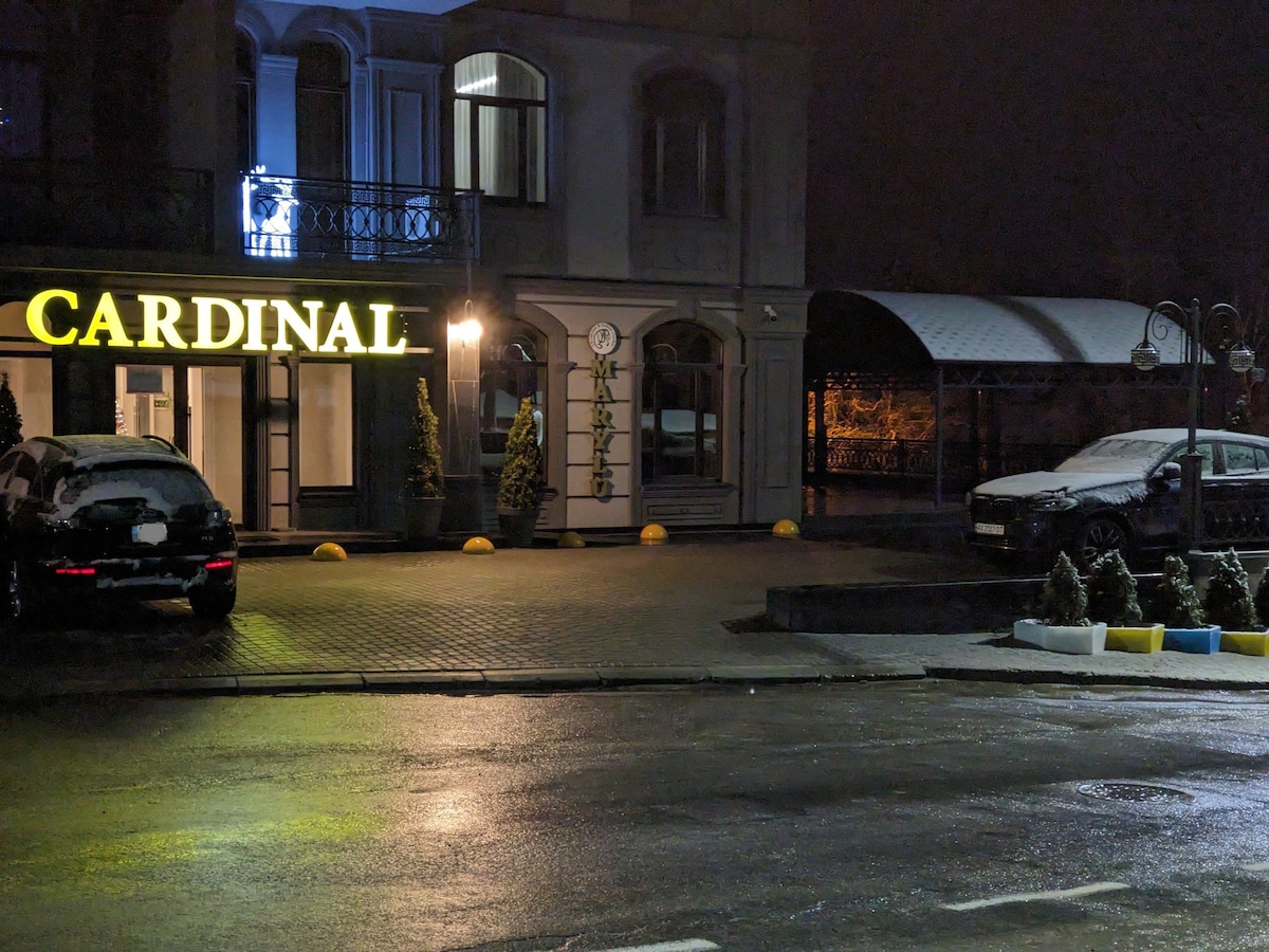 The exterior view of the CARDINAL aparthotel is illuminated at night, featuring the hotel's name prominently displayed. A well-maintained parking area is visible, with several vehicles parked, and decorative greenery enhances the welcoming entrance. Soft lighting provides a serene ambience.