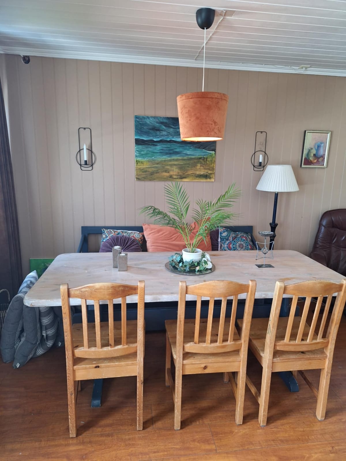 A dining area features a wooden table with seating for six, complemented by a child’s high chair. A potted plant and decorative elements adorn the table, while a warm light fixture hangs above. The walls display art and soft furnishings, creating a welcoming space.
