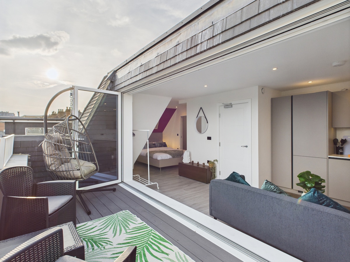 A spacious private balcony features a stylish hanging chair and modern furnishings. The surrounding area is complemented by light wood decking and lush green accents. Large sliding doors open into a bright living space, showcasing a cozy bedroom in the background.