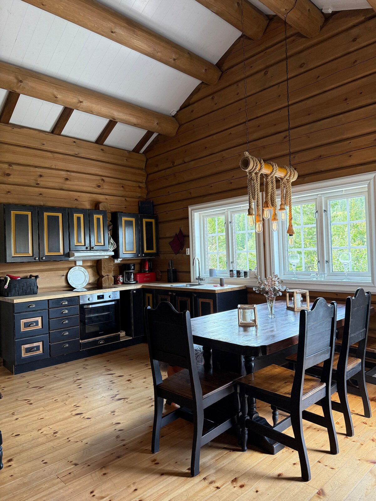 A spacious kitchen features wooden beams and walls, accented by dark cabinetry with gold detailing. A large dining table is surrounded by six chairs, and large windows allow natural light to illuminate the area. Kitchen appliances and utensils are neatly arranged.