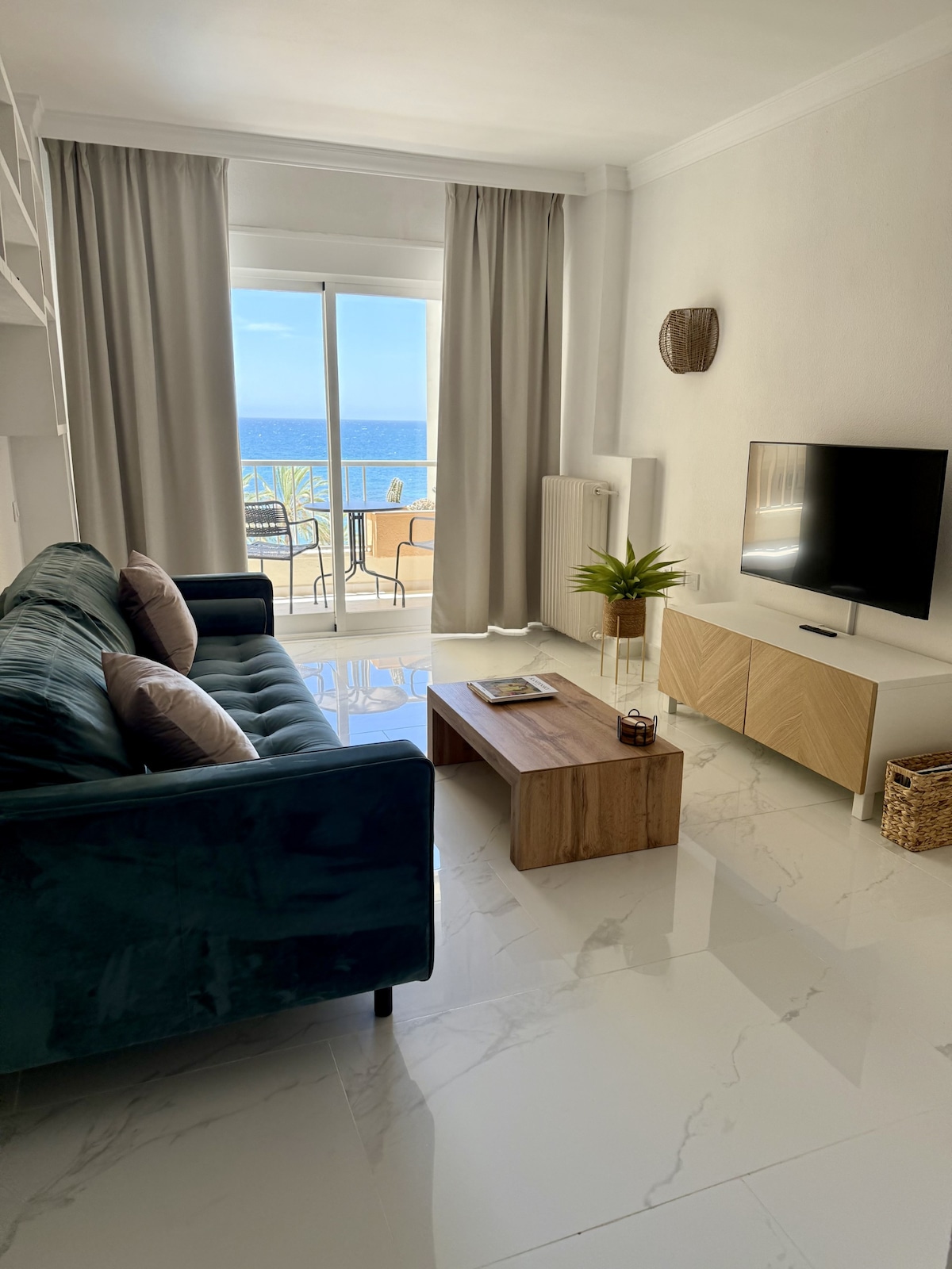 A bright living area features a teal sofa with decorative pillows and a wooden coffee table. A flat-screen TV is mounted on a light wood entertainment unit. Large glass doors open to a balcony with sea views, framed by soft curtains.