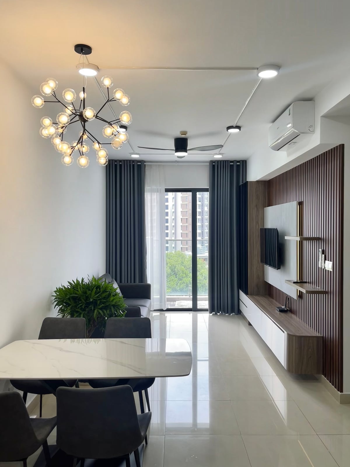 A modern living area features a stylish lighting fixture above a white dining table surrounded by gray chairs. A wall-mounted television is displayed, with a wooden accent wall as a backdrop. Large windows allow natural light to enter, complemented by sheer curtains.
