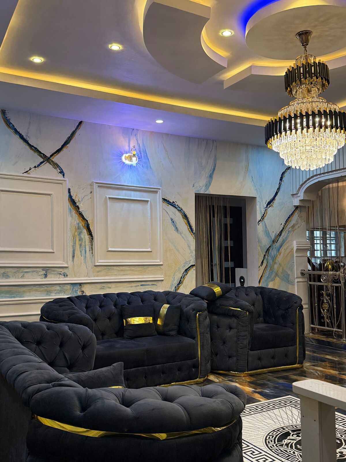 The living area features an elegant chandelier hanging from a decorative ceiling. Plush, dark blue sofas with gold accents are arranged to encourage conversation. Soft lighting highlights the marble-inspired wall design, creating a sophisticated and inviting space.