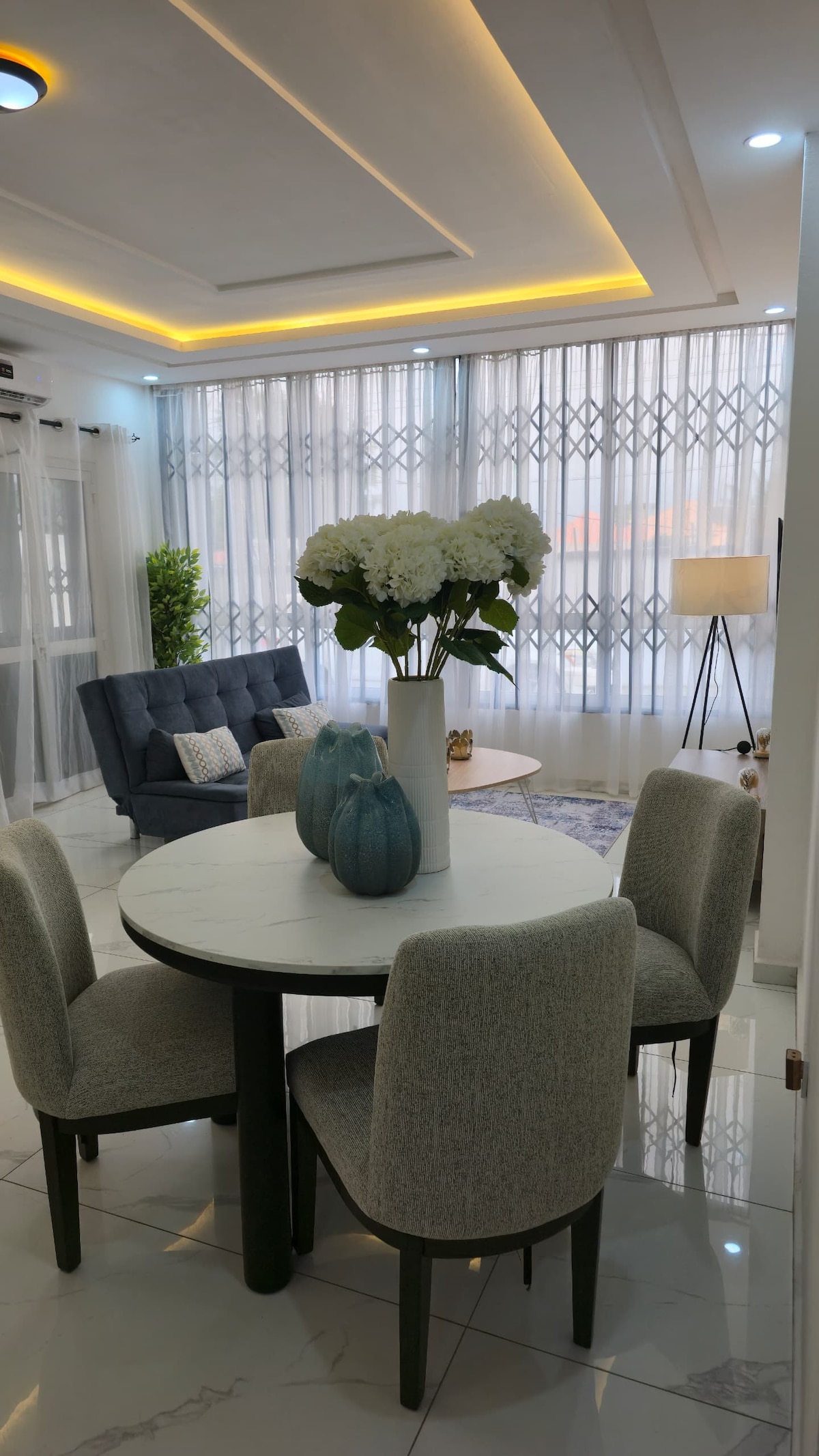 A modern dining area features a round marble table surrounded by four upholstered chairs. A tall vase with white flowers adds a natural touch. The spacious living area includes a blue sofa and a large window adorned with sheer curtains, allowing ample natural light.