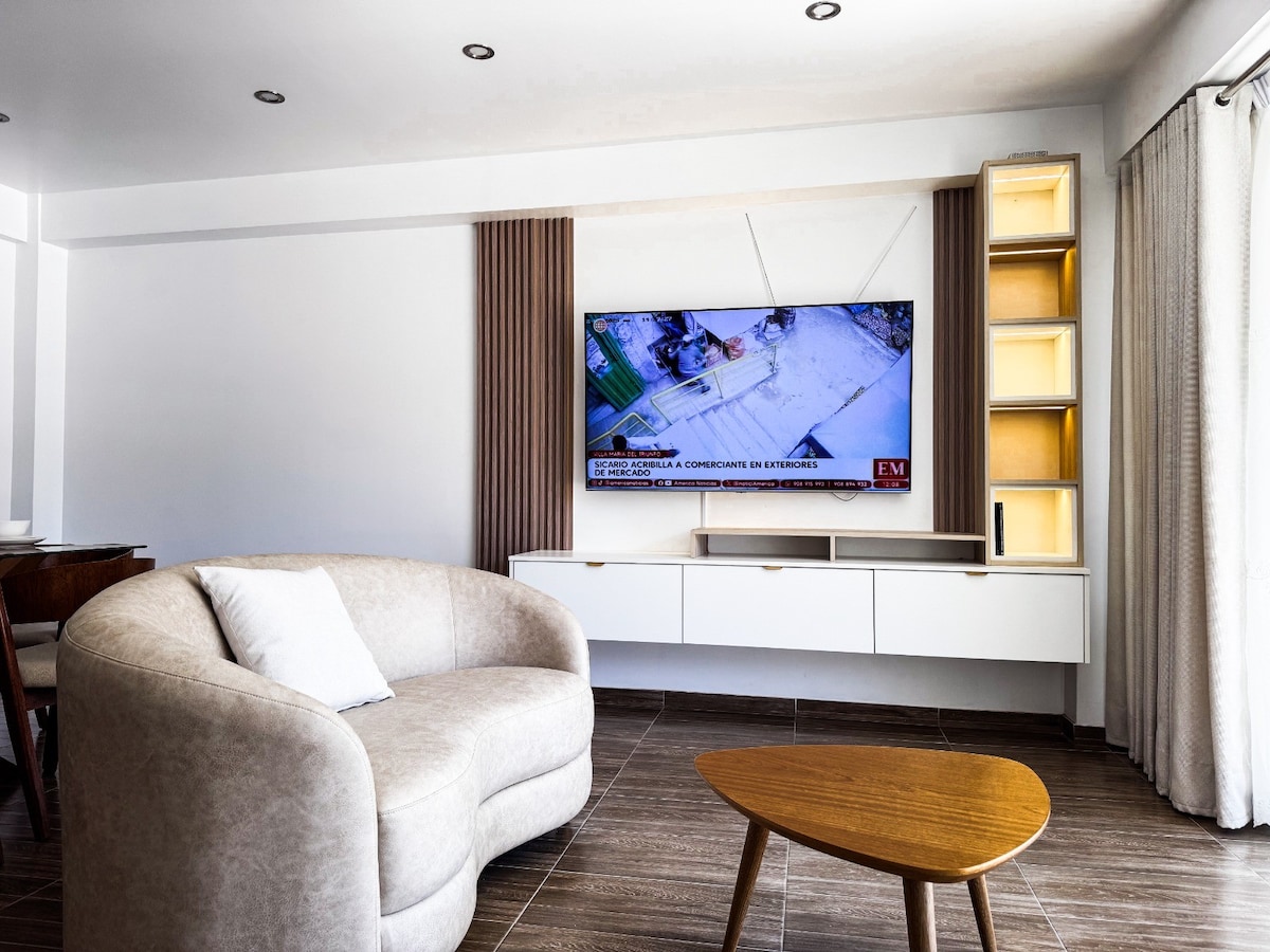 A modern living area is highlighted by a curved light-colored sofa, positioned across from a mounted television. A minimalist wooden coffee table offers a functional accent, while soft beige curtains frame the large window, allowing natural light to brighten the space.