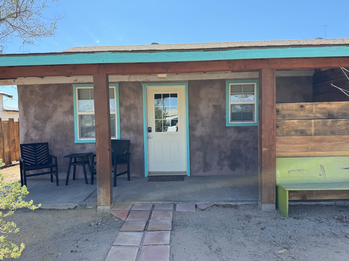 The entrance to the apartment features a welcoming porch area with two black chairs positioned beside a vibrant green bench. A light blue trim outlines the door and windows, while the textured exterior wall exhibits a warm earth tone. A clear blue sky is visible above.