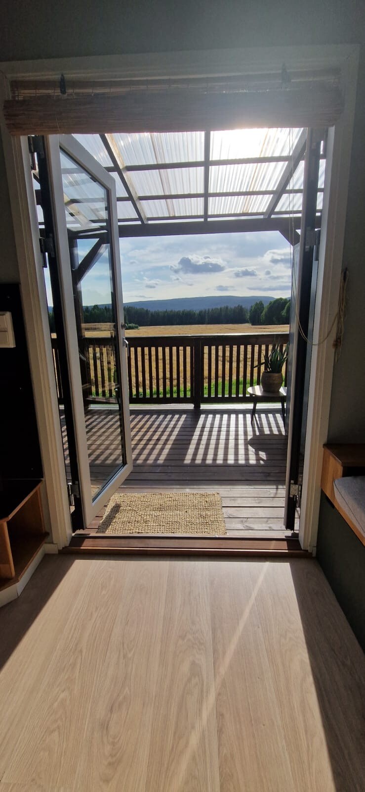 A view from the entryway features glass doors opening to a spacious terrace. Sunlight filters through the polycarbonate roofing, illuminating the interior. The scene beyond presents a wide, open landscape with greenery and clouds in the distance.