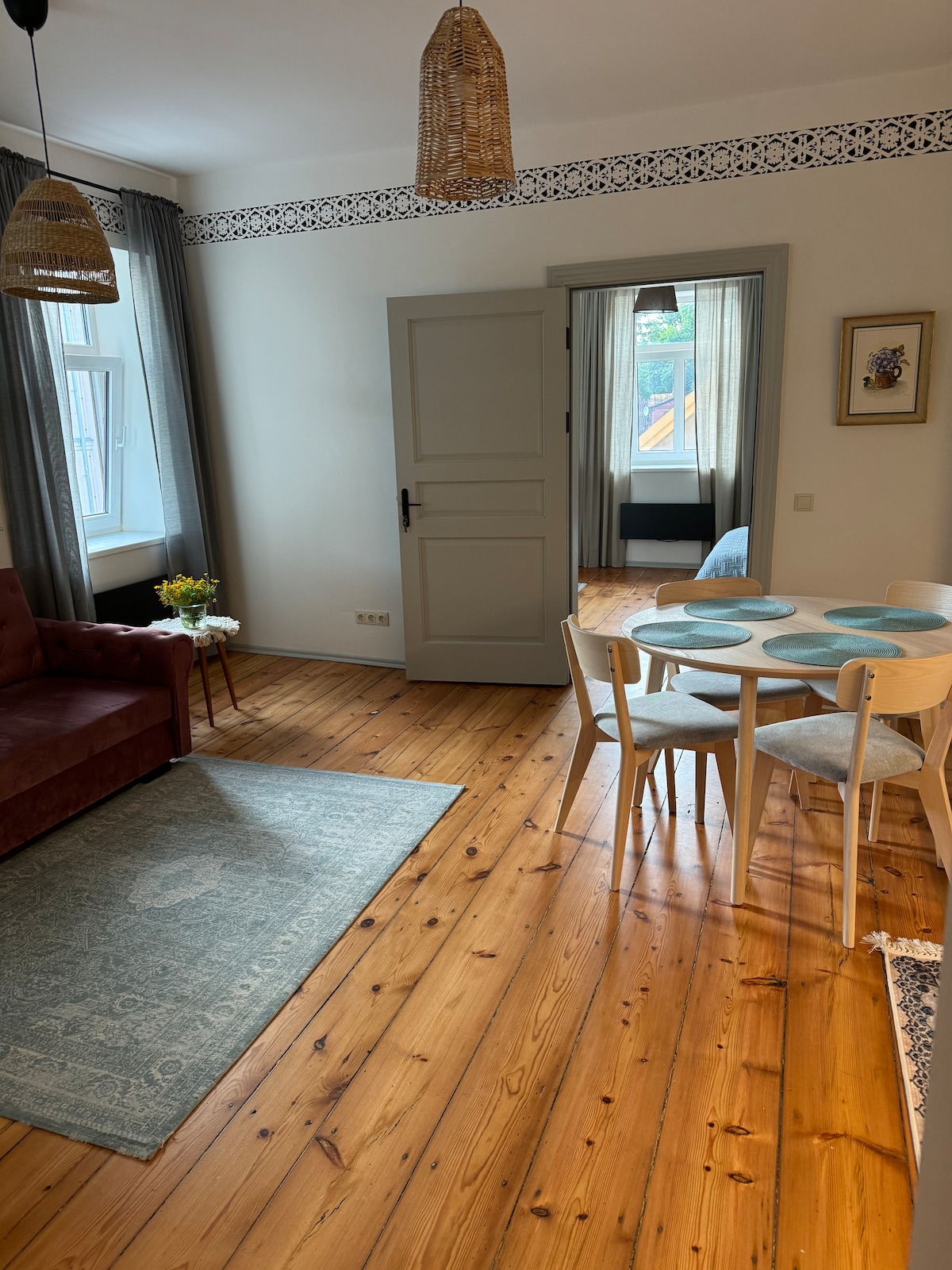 A welcoming living space features a deep red couch, a small dining table with light-colored chairs, and a blue area rug on wooden flooring. Natural light filters through two windows, and decor elements include woven light fixtures and a framed artwork on the wall.