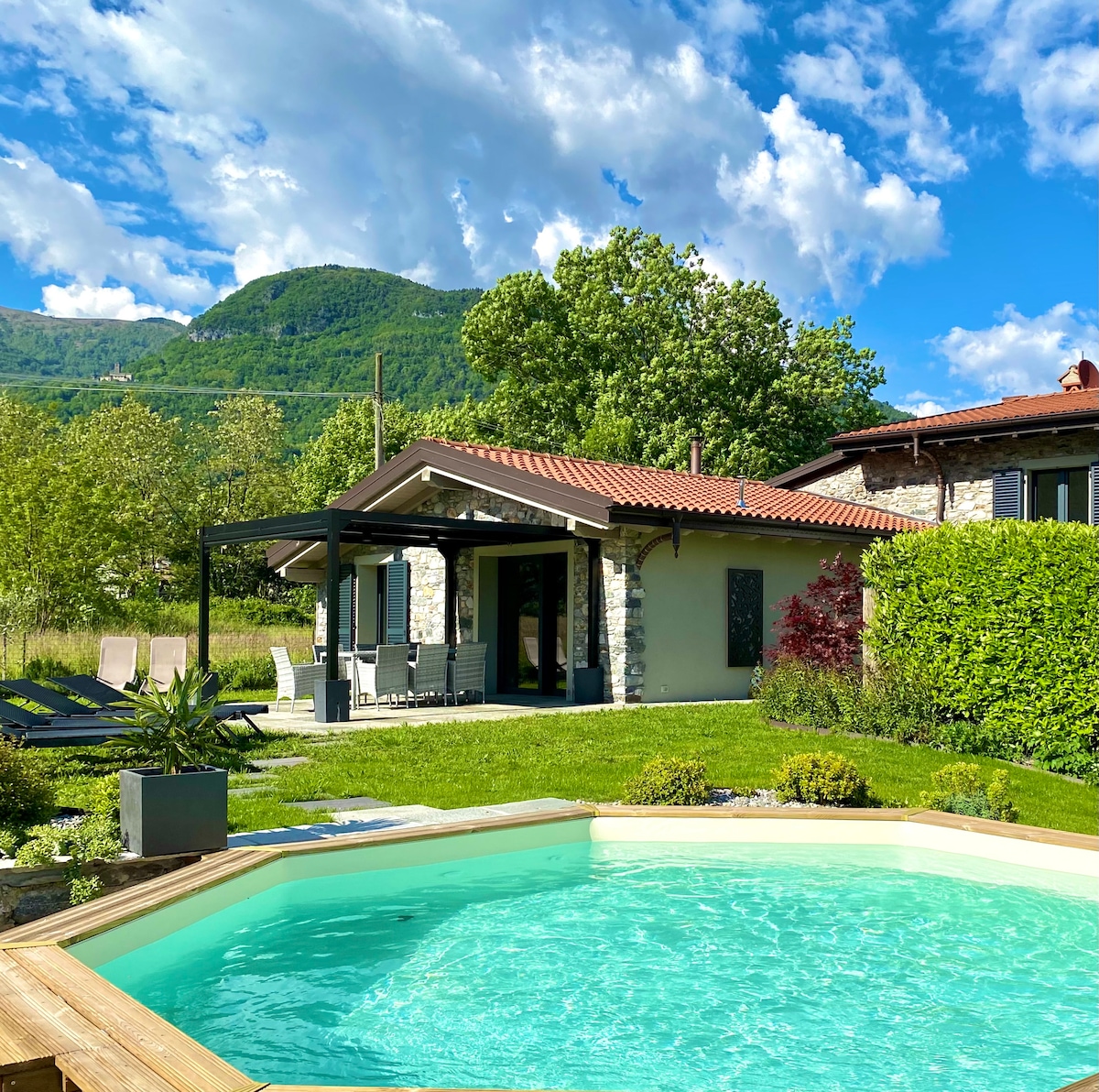 The image captures a sparkling private pool surrounded by a well-maintained garden. In the background, a covered patio with outdoor seating is visible, and lush green mountains dot the skyline under a partly cloudy blue sky.