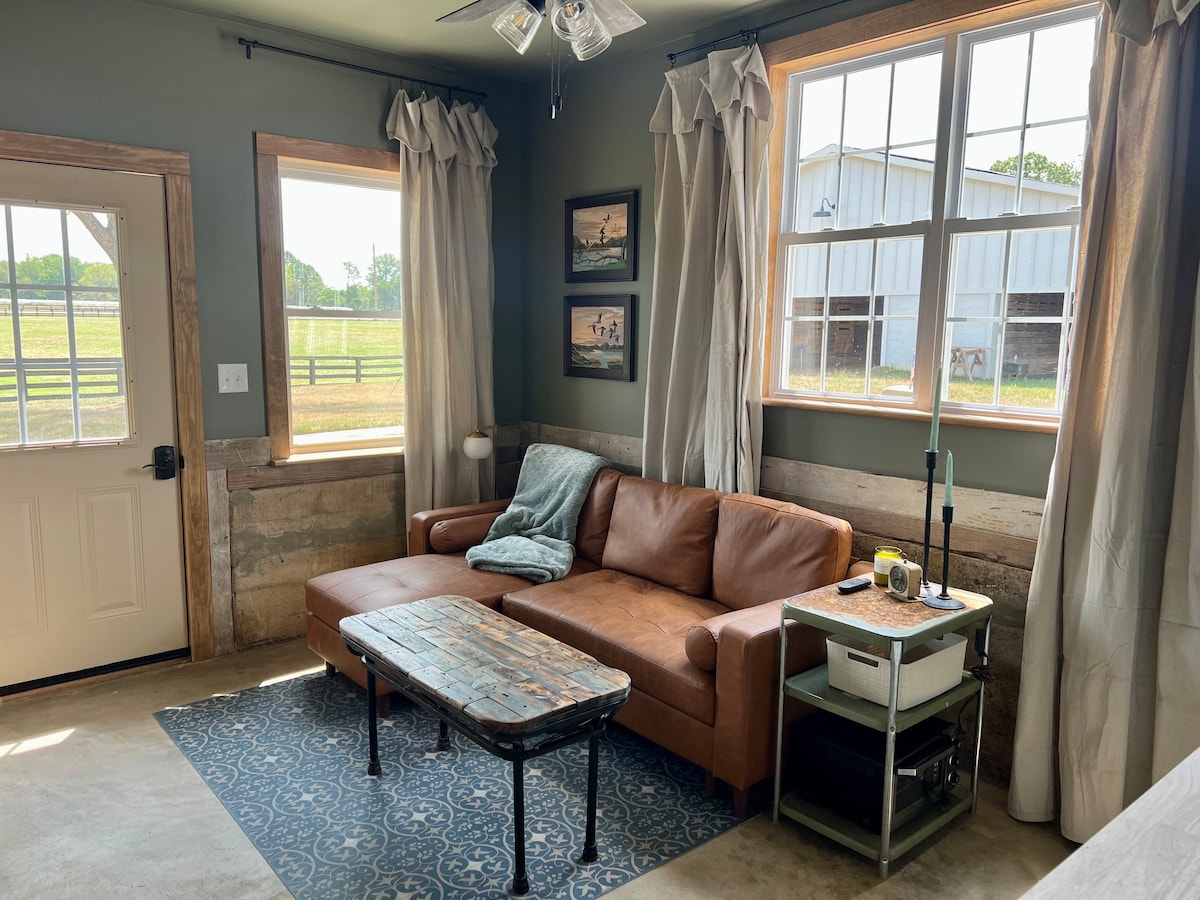 A cozy living room features a leather sectional couch with a soft throw blanket draped over it. A rustic coffee table sits in front, while a small side table is beside the couch. Large windows provide natural light and a view of the farm outside.