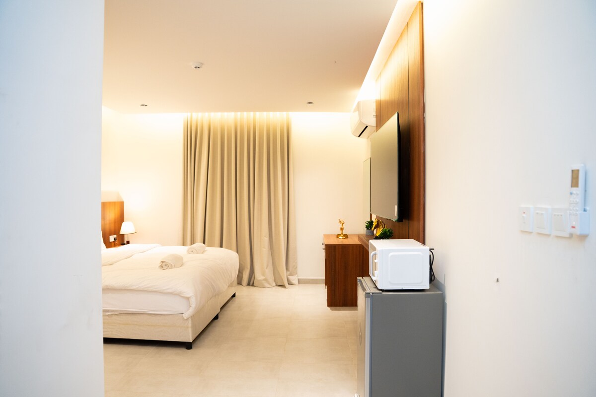 A serene bedroom is displayed, featuring a spacious bed with neutral bedding and two towels neatly arranged. A wall-mounted television is present above a wooden cabinet, while soft lighting is diffused through light curtains. A microwave and mini-fridge are positioned beside the entrance.