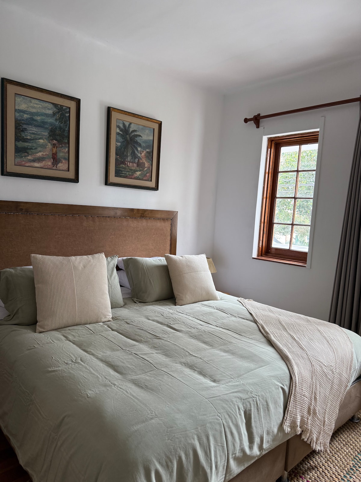 A spacious sleeping area features a comfortable bed with soft bedding and an elegantly draped throw. Two framed artworks adorn the wall above the bed. A wooden window allows natural light to fill the room, complemented by a neutral-toned curtain.