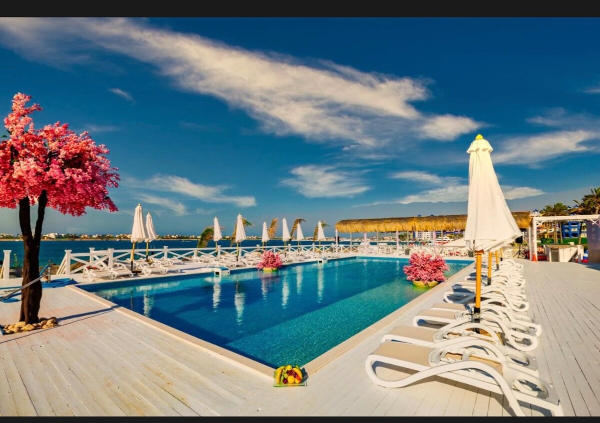 A serene pool area is surrounded by white lounge chairs and vibrant pink flowering trees. The clear blue water reflects the sky, while several umbrellas provide shade. The scene is complemented by a wooden deck, creating a relaxing atmosphere for guests.