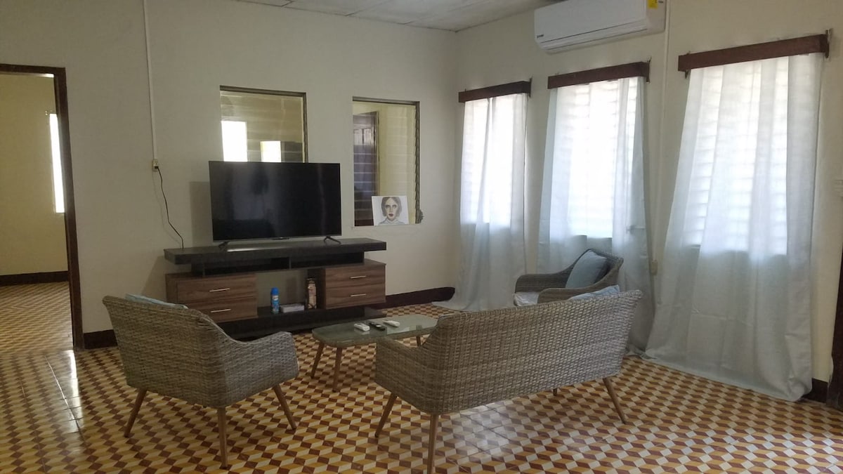 A living area is presented with tiled flooring and a neutral color scheme. A large television is mounted on the wall above a low-profile media console. Two patterned armchairs are placed beside a glass coffee table, with natural light filtering through sheer white curtains on the windows.