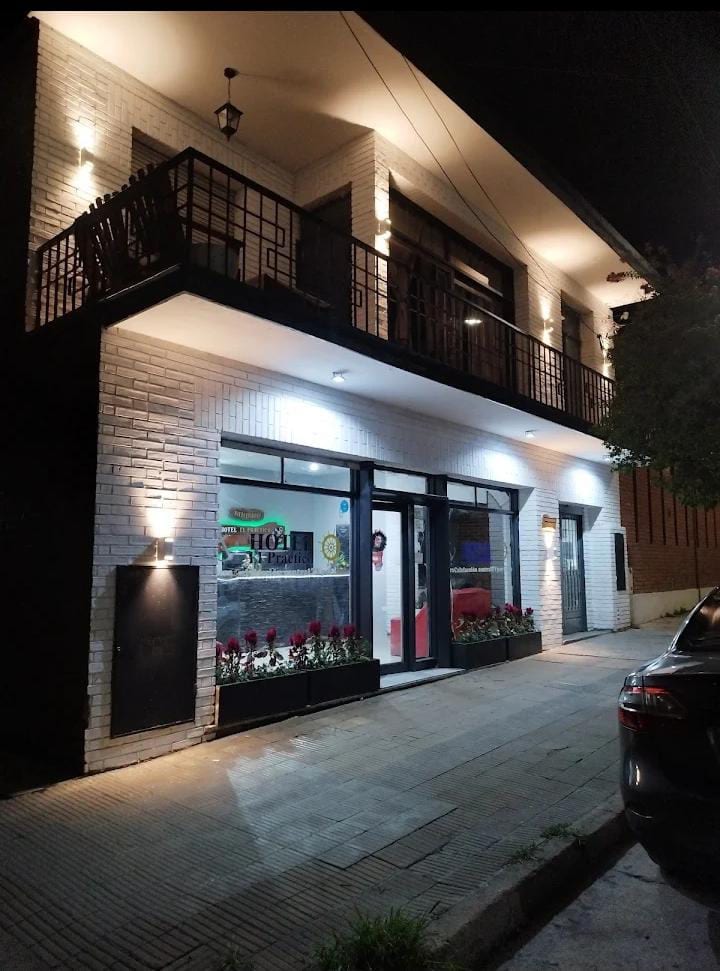 The exterior of the building is shown at night, featuring a two-story structure with a modern facade. Warm lighting illuminates the entrance and balcony. Flower beds line the ground floor windows, enhancing the welcoming appearance of the space.