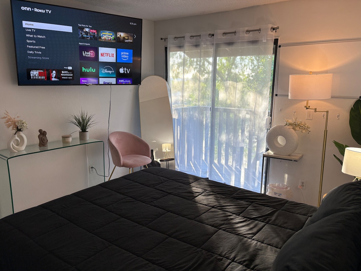 A comfortable bedroom features a spacious bed covered with a dark quilt. A large flat-screen TV is mounted on the wall, displaying various streaming options. Natural light enters through glass doors leading to a small balcony. A modern white lamp and a round mirror complement the decor.