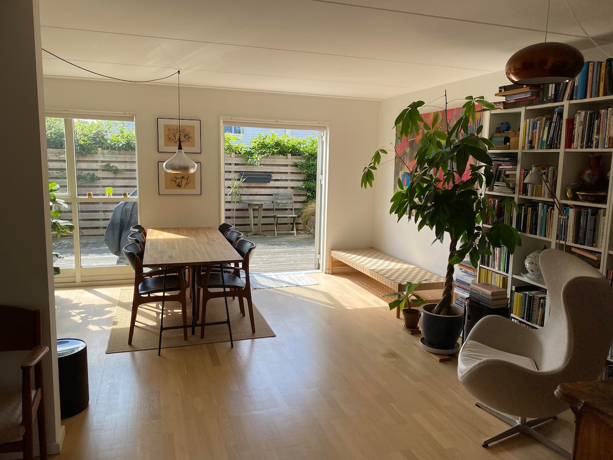 The living area features a dining table surrounded by six chairs, situated near large sliding glass doors that open to an outdoor space. A potted plant adds greenery, while a bookshelf filled with books is positioned against one wall. Natural light illuminates the room.