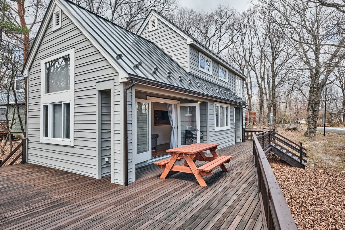A spacious wooden deck extends from the house, surrounded by trees. A picnic table with attached benches is positioned on the deck, inviting outdoor dining. The structure features a combination of large windows and a distinctive roof design, showcasing a modern country style.