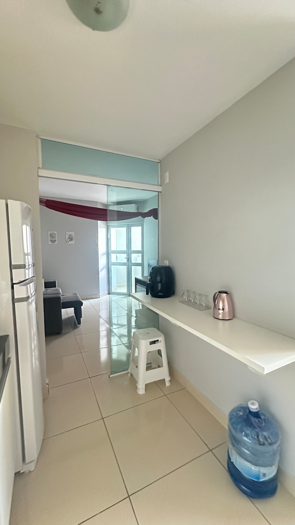 A sleek hallway connects a modern kitchen area to a cozy living space. A white countertop is adorned with a coffee maker and a rose gold kettle, while a water dispenser stands to the side. Light enters through a glass door, enhancing the airy feel.