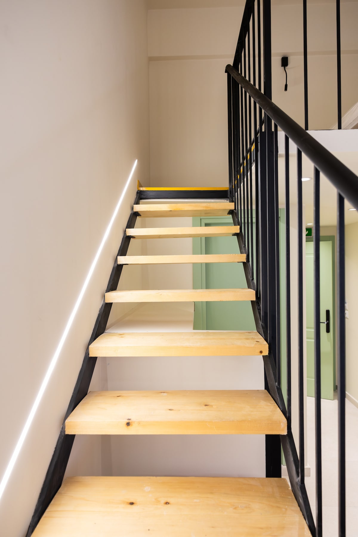 A set of wooden stairs features a simple design with open risers and a sleek black railing. Soft lighting is incorporated along the edge of the steps, guiding upward towards an upper level.