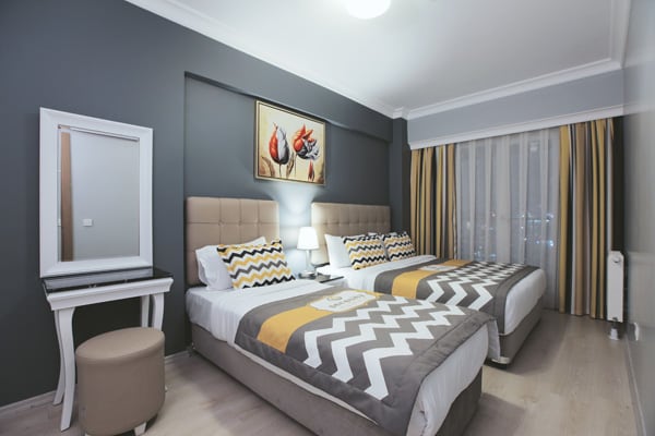 A modern guestroom features two spacious beds with patterned bedding in neutral tones accented by vibrant yellow. A small white desk and a round stool are positioned beside the beds. Natural light filters through sheer curtains, enhancing the calming atmosphere.