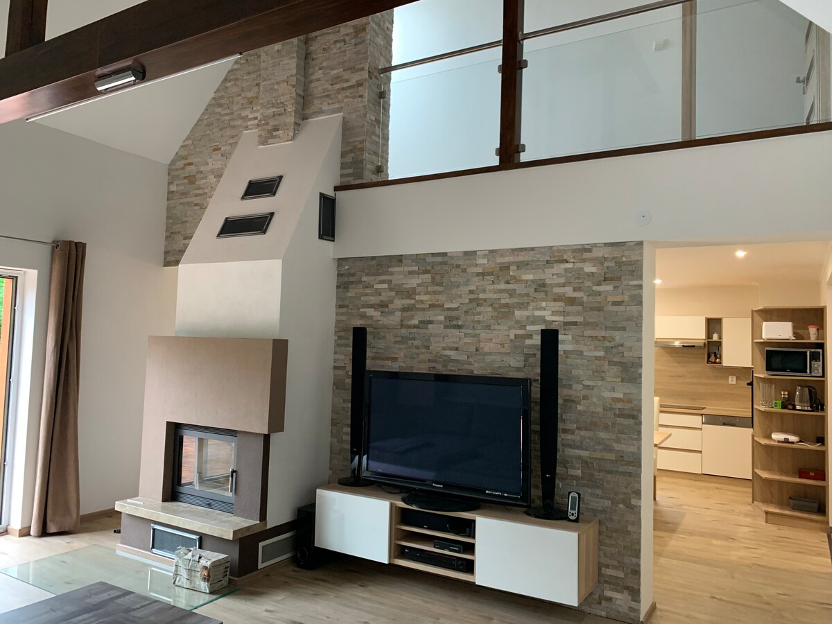 A modern living area features a prominent stone fireplace and a sleek television mounted on the wall. Natural light is welcomed through large windows, with a view of the open kitchen area in the background, creating an inviting and spacious environment.