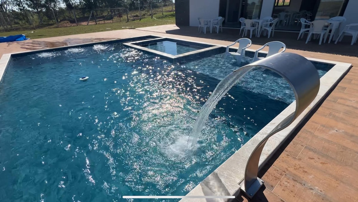 A swimming pool is showcased with a modern water feature, creating a gentle cascade. The sun reflects off the clear blue water, while white lounge chairs are positioned nearby on the tiled deck, offering a comfortable space for relaxation.