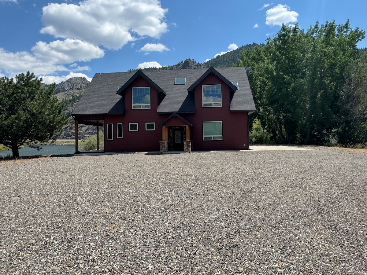 The exterior of a spacious lodge is visible, featuring a red wood façade and multiple windows. A gravel driveway leads to the entrance. Surrounding trees and a mountainous backdrop complement the scenic location near the Missouri River, creating an inviting outdoor space.