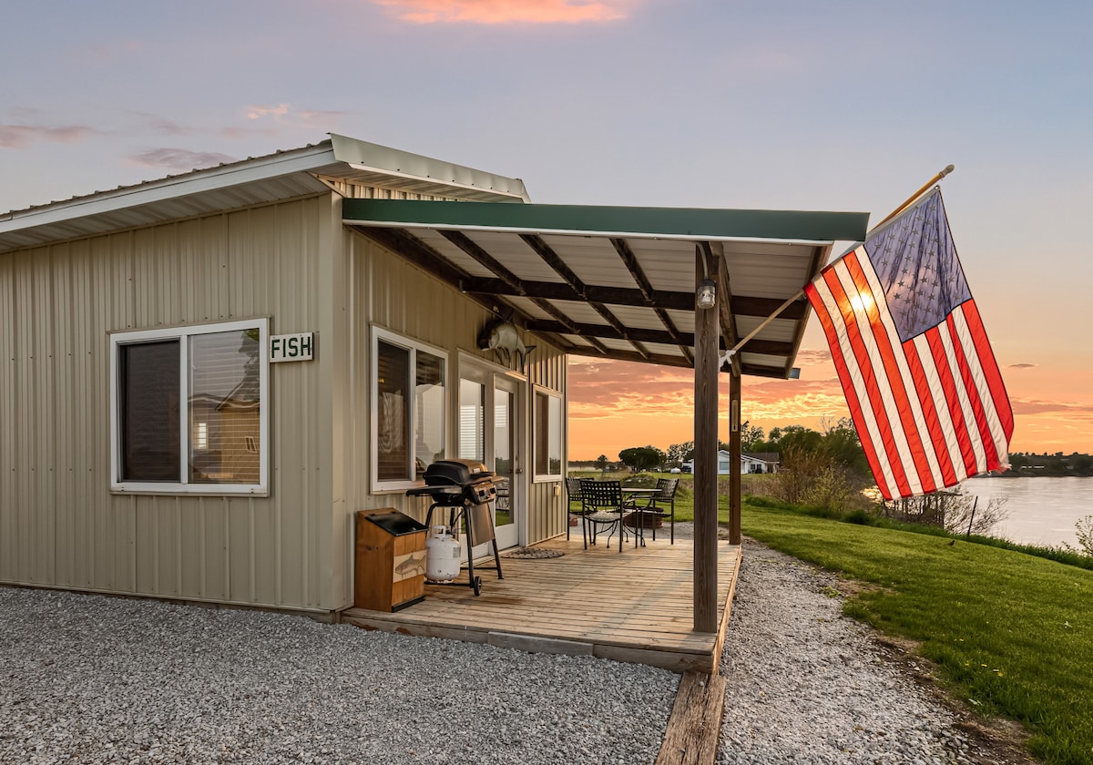 A cabin exterior features a covered porch with a wooden deck, where a grill, dining set, and an American flag are prominently displayed. The backdrop includes a serene view of water and greenery, illuminated by a soft sunset glow.