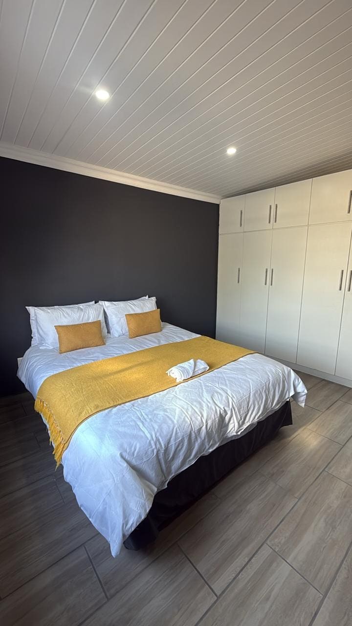 A comfortable bedroom is presented with a spacious bed adorned with a white quilt and a yellow throw. Two accent pillows complement the bedding. The room features light-colored cabinetry and is illuminated by recessed ceiling lights, enhancing its cozy ambiance.