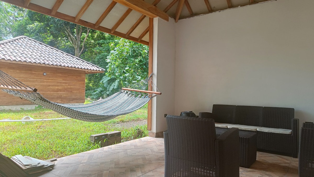 A covered outdoor seating area is featured, with a hammock suspended nearby. Comfortable wicker furniture includes a sofa and chairs arranged around a wooden coffee table. Lush greenery surrounds the space, and a wooden building is visible in the background.