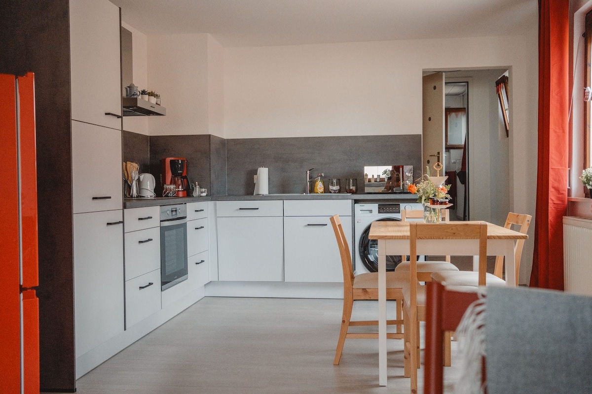 A modern kitchen features sleek white cabinetry and a dark backsplash. A wooden table with four chairs sits in the foreground, while appliances and kitchenware are neatly arranged on the counter. Natural light enters through a window adorned with red curtains.