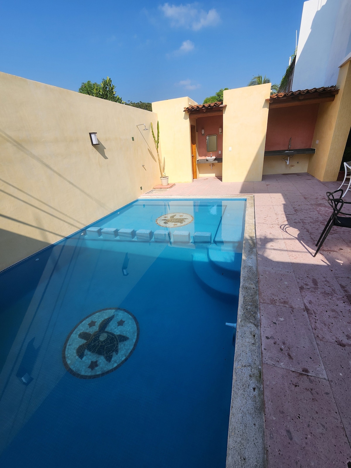 A serene pool area is depicted, featuring clear blue water with a decorative tile design at the bottom. Surrounding walls are painted a warm yellow, and there is a shaded seating area with a chair. A nearby outdoor shower and sink are visible.