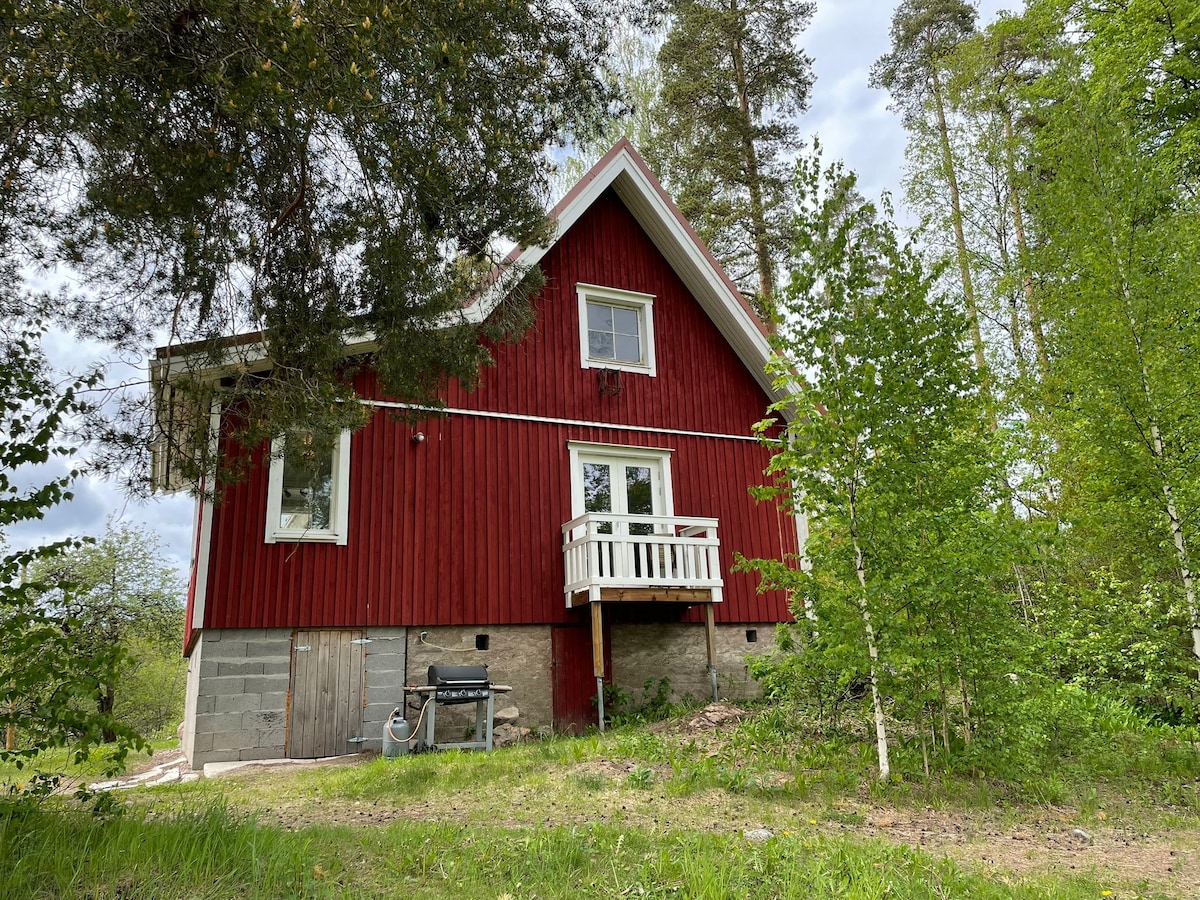 A charming red house is framed by greenery, featuring a white balcony and a ground-floor entrance. The home is surrounded by trees and grass, creating a natural, serene environment. A barbecue grill is placed nearby, indicating outdoor cooking possibilities.