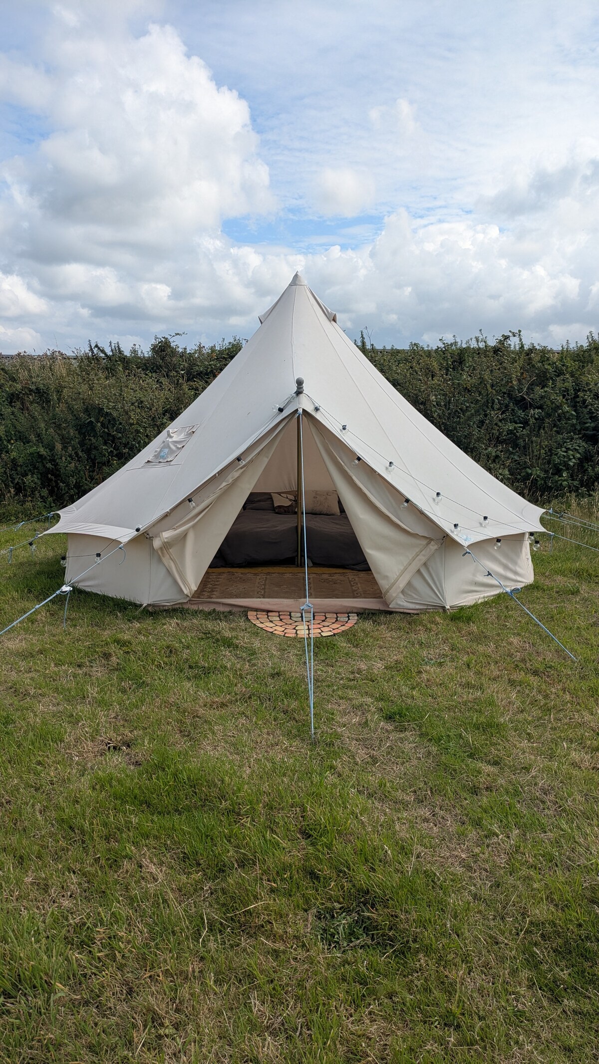 A spacious bell tent is displayed, featuring a cream-colored exterior with a conical shape. It is positioned in a grassy area surrounded by low vegetation. Tent flaps are open, revealing a glimpse of the interior. A circular mat is placed at the entrance.