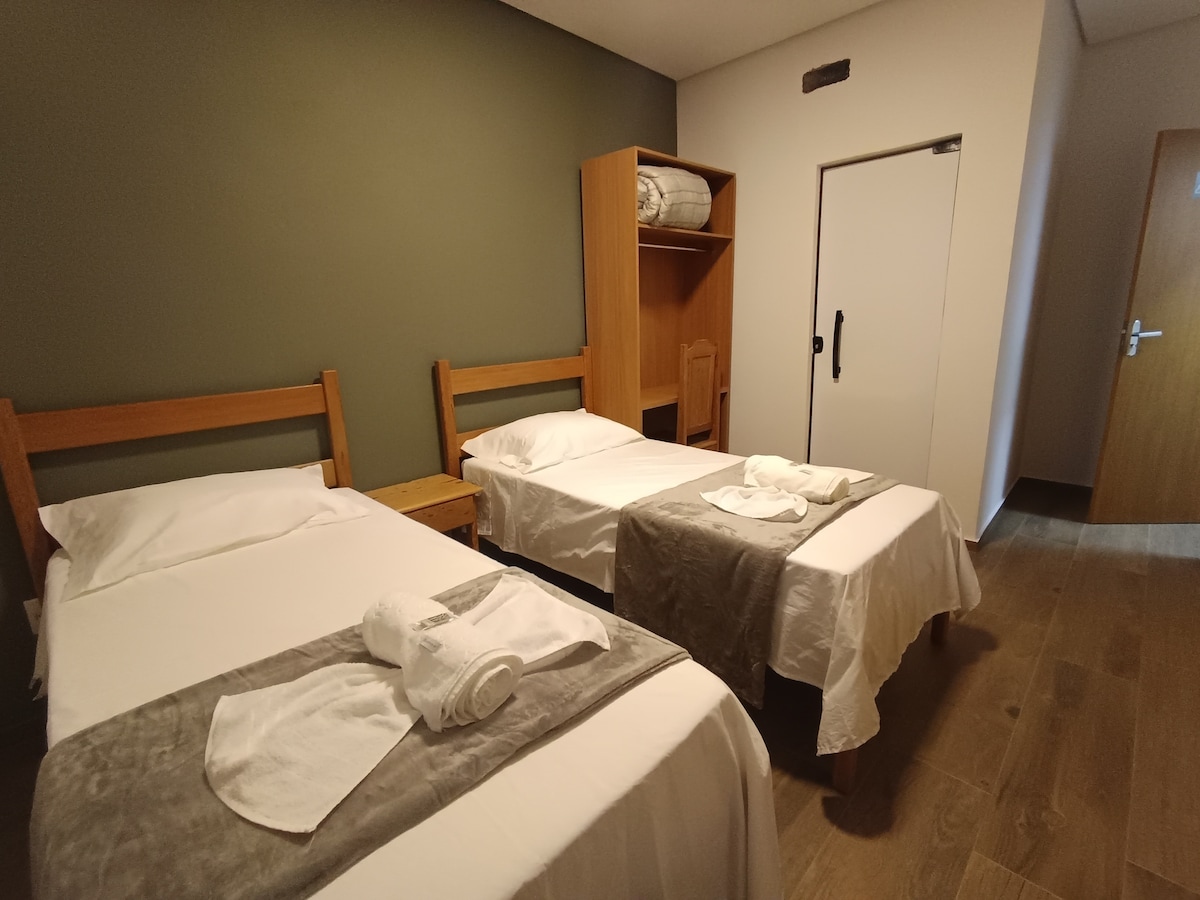 The room contains two twin beds, each dressed in soft linens. A muted green wall serves as a backdrop, while wood furniture, including a nightstand and shelving unit, adds warmth. Light enters through a nearby door, enhancing the room's spacious feel.