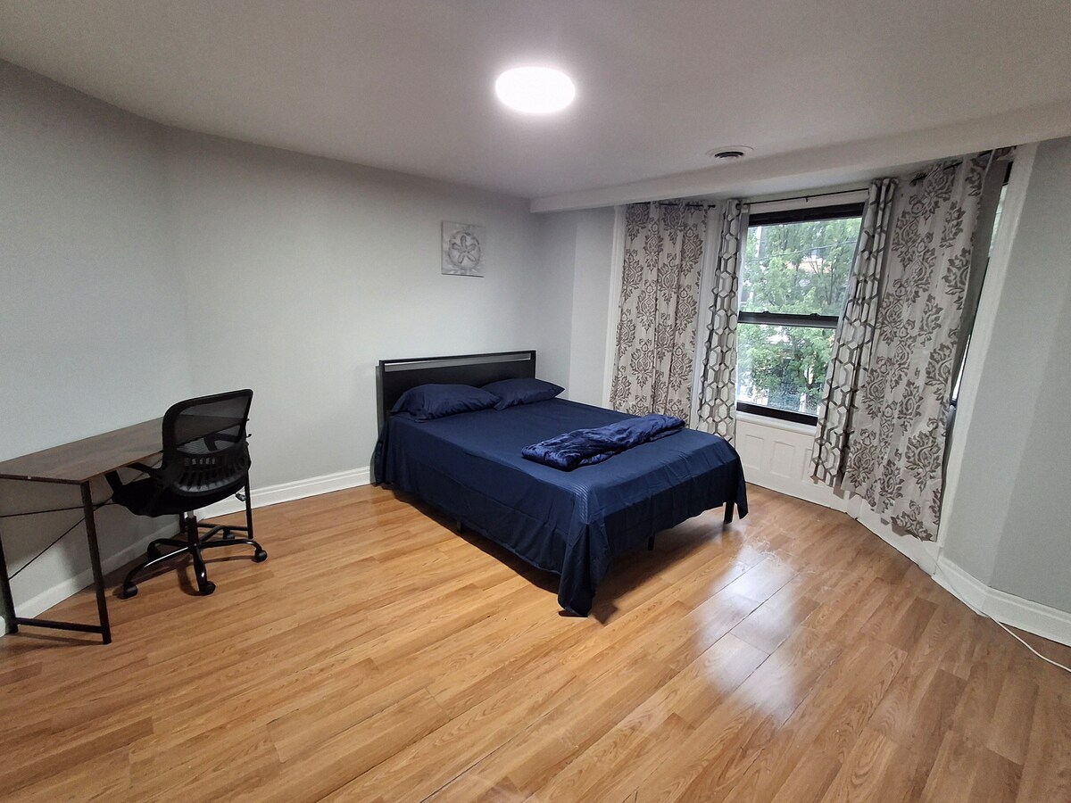 A spacious bedroom features a large bed with dark blue linens. A desk and chair are positioned nearby, while large windows allow natural light to fill the room. Light-colored curtains complement the neutral walls, creating a calming environment.