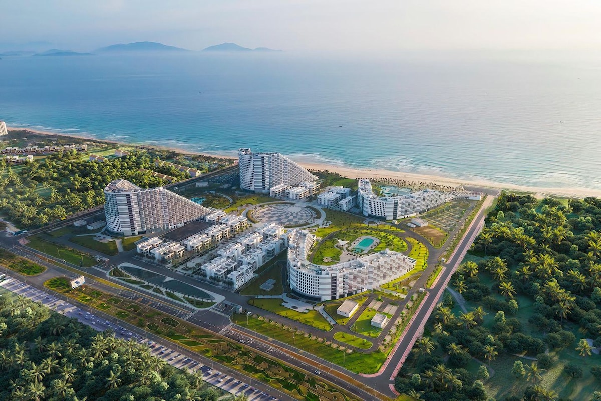 An aerial view captures a sprawling resort complex nestled among palm trees, with modern buildings surrounding landscaped gardens. The coastline stretches along the horizon, showcasing the tranquil ocean waters, while pathways and pool areas are visible throughout the property.