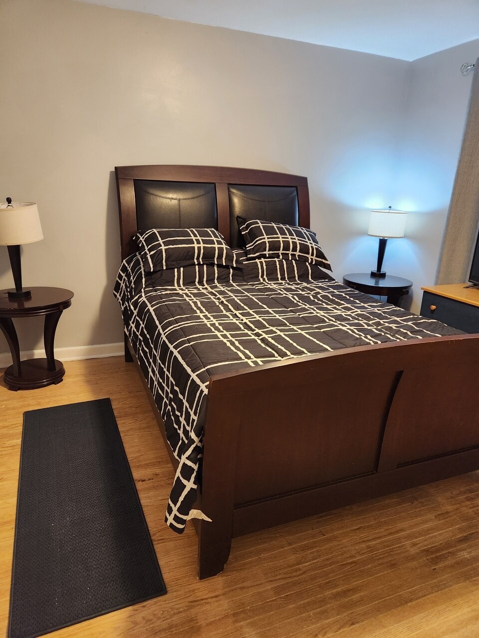A comfortable bedroom features a large bed with a modern patterned duvet cover. Bedside tables are placed on either side, each equipped with a lamp for evening illumination. Natural light filters in from a window, adding to the room's relaxed ambiance.