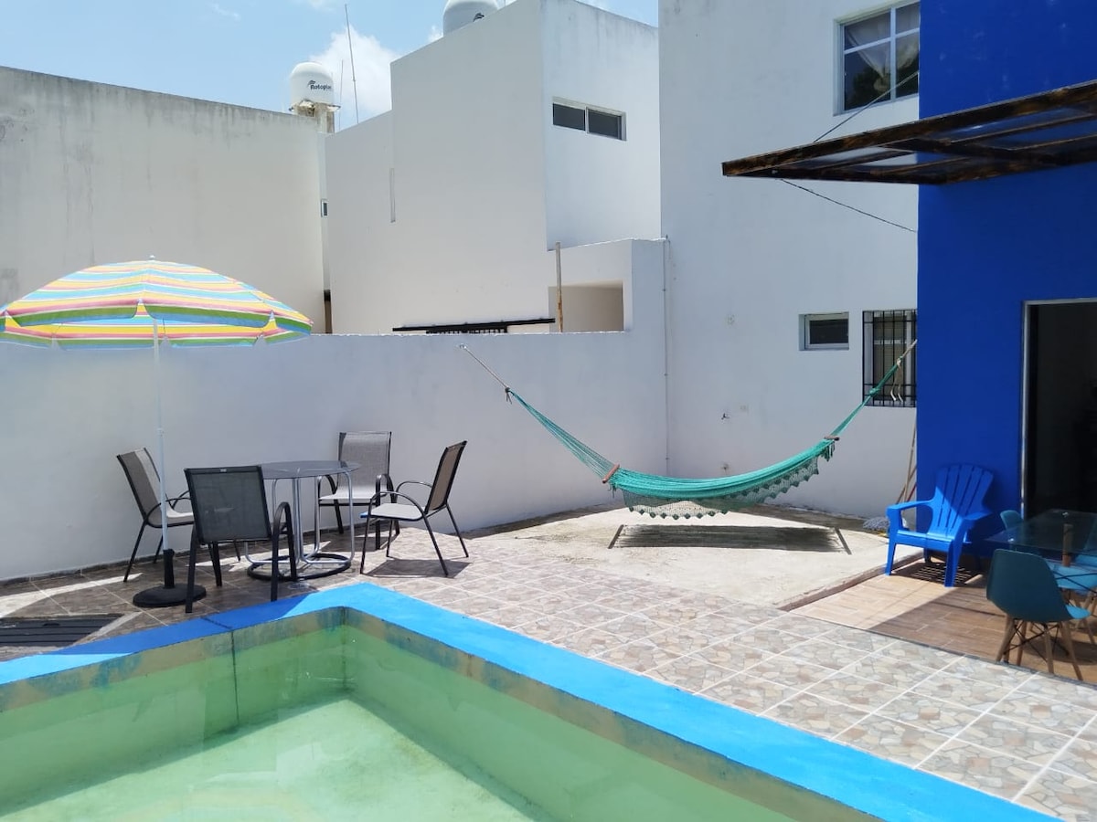 A private outdoor space is featured, showcasing a turquoise pool surrounded by patterned tiles. A shaded table with four chairs is positioned nearby, along with a hammock suspended between two structures. An umbrella provides additional shade, enhancing the relaxation area.